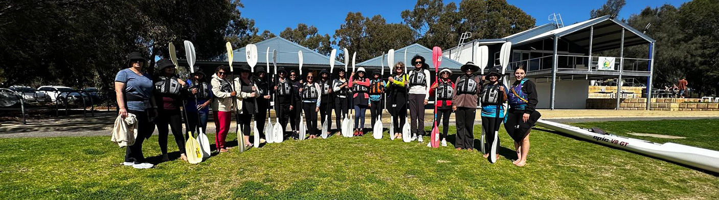 A group of paddlers stand in front of a boat shed with oars and wearing flotation vests