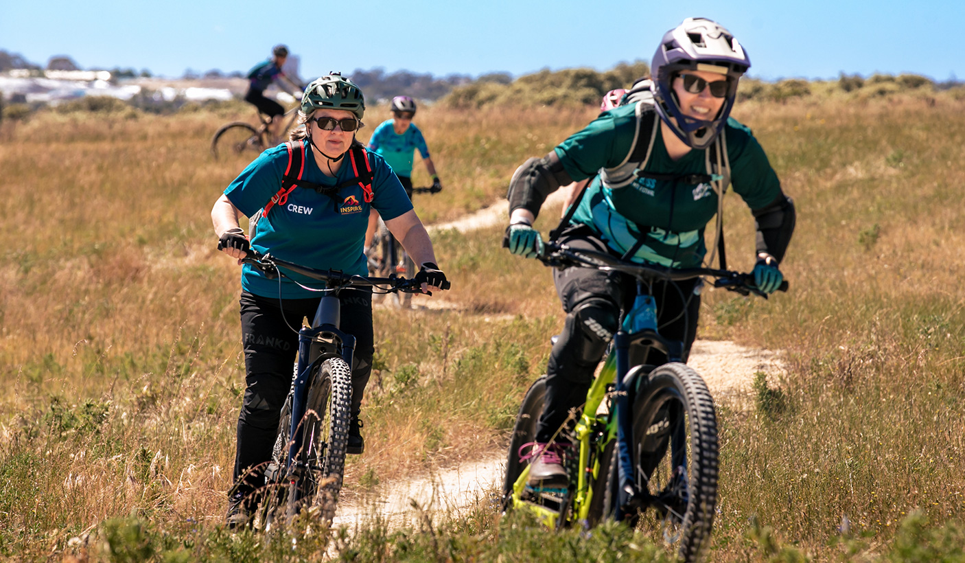 A small group of women ride mountain bikes on a path, in a grassy landscape.