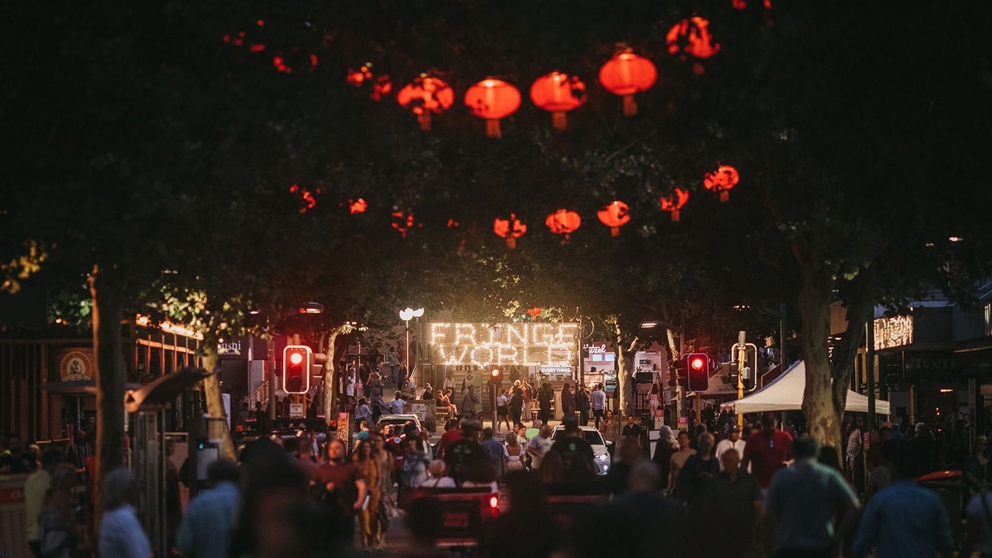 A night time image of people in the street, with lanterns hanging over the street and an illuminated sign reads 