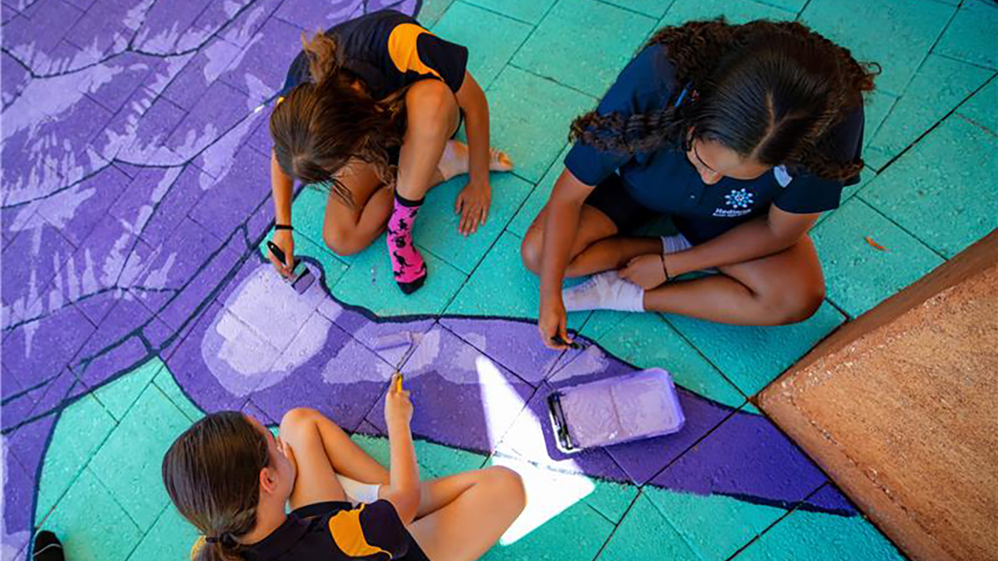 3 school students sit on a paved area painting a colourful floor mural