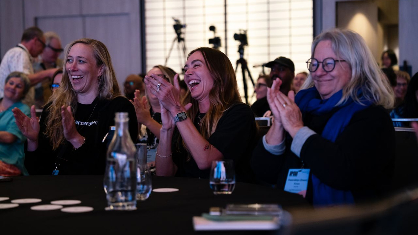 Three people sitting at a table, at a conference, laugh and clap