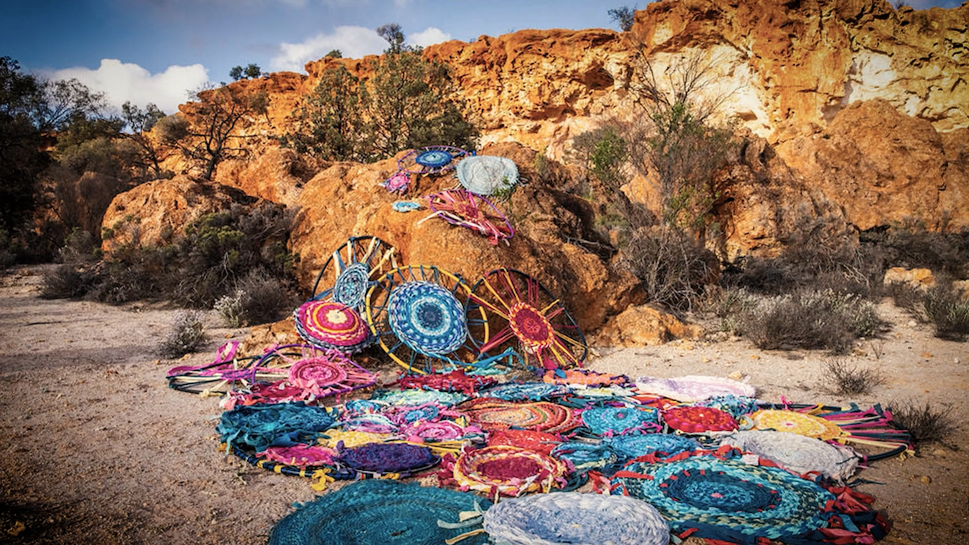 A number of weavings spread out over the ground in a arid rocky landscape.