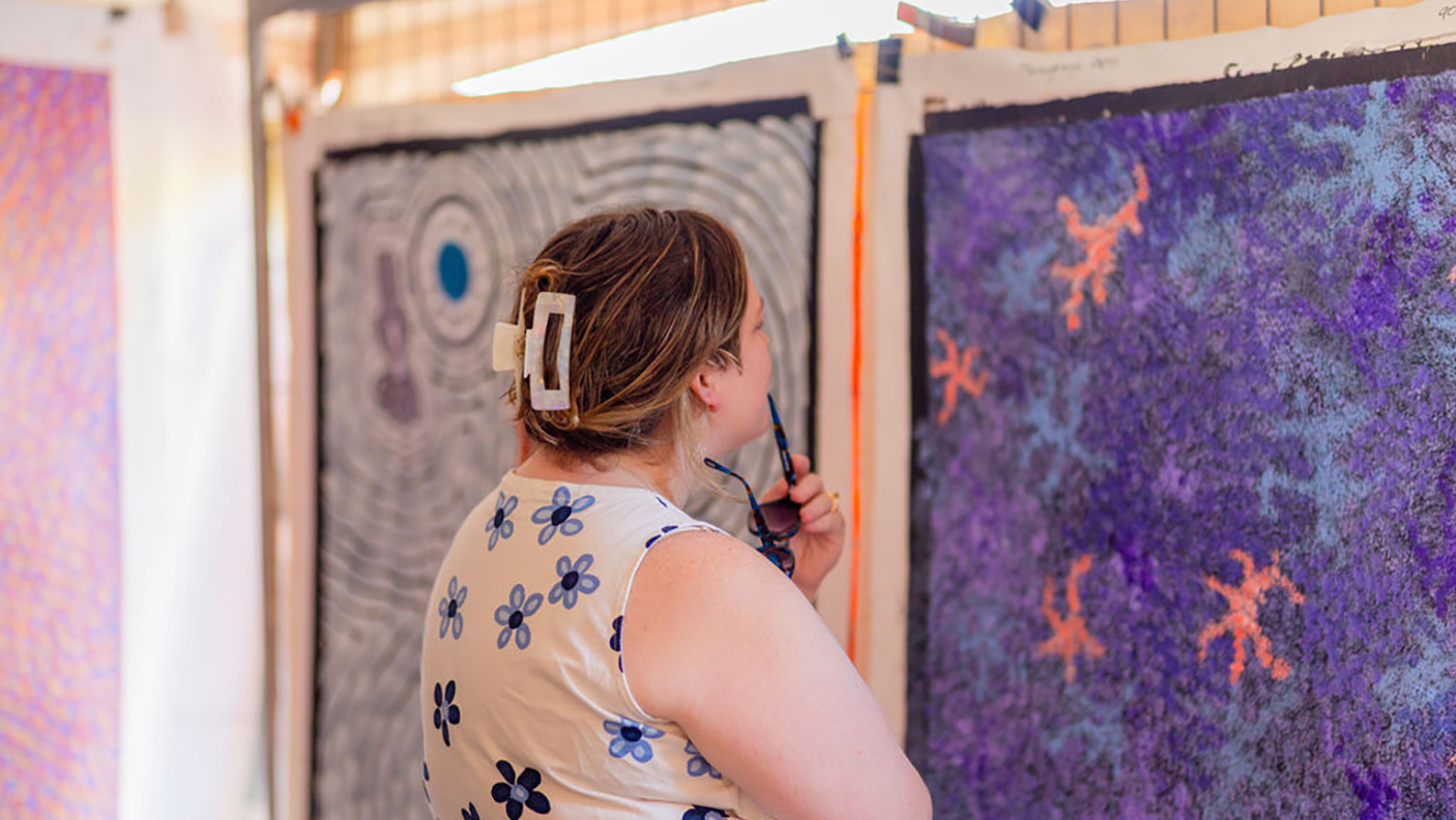 A woman looks at unframed painting on canvas, hanging in a market stall
