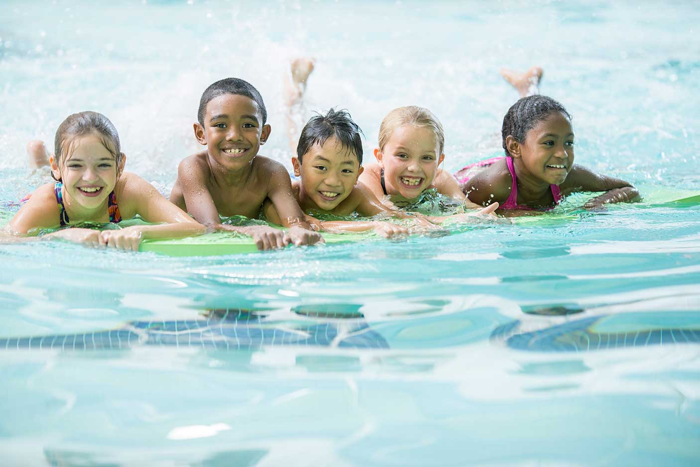 A multi-ethnic group of elementary age children are swimming on their kick boards during a swimming lesson. They are smiling and looking at the camera.