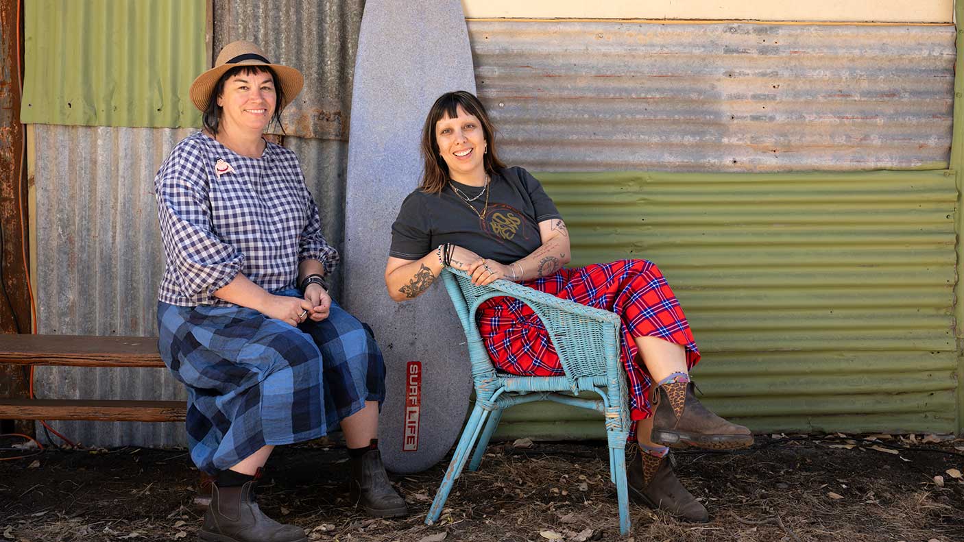 Two women sitting down in front of a corrugated wall with a surf board standing up against it,