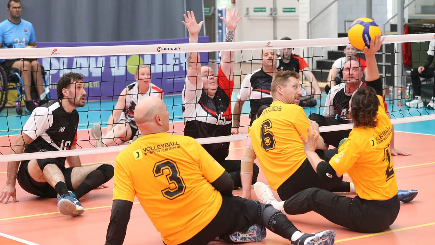 two teams of volleyball players sit on an indoor court, playing adaptive volleyball
