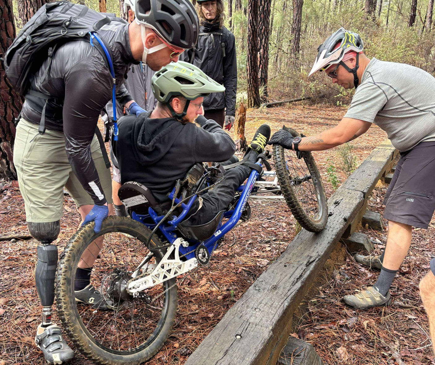 a man on an adaptive mountain bike is assisted on a trail by able bodied people.