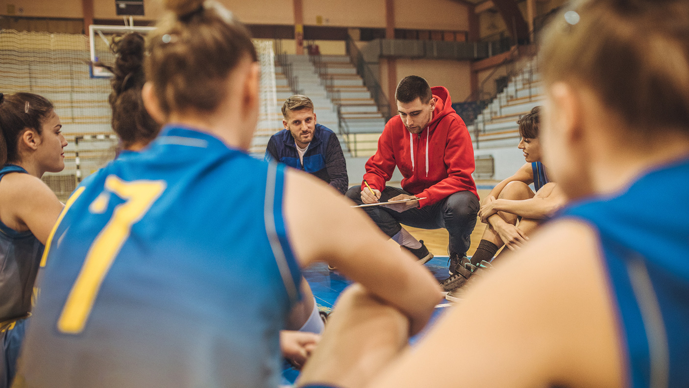 A coach huddles around a women's sport team on an indoor court