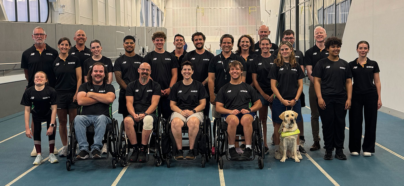 A group of 22 athletes and a guide dog pose for the camera on an indoor sports court