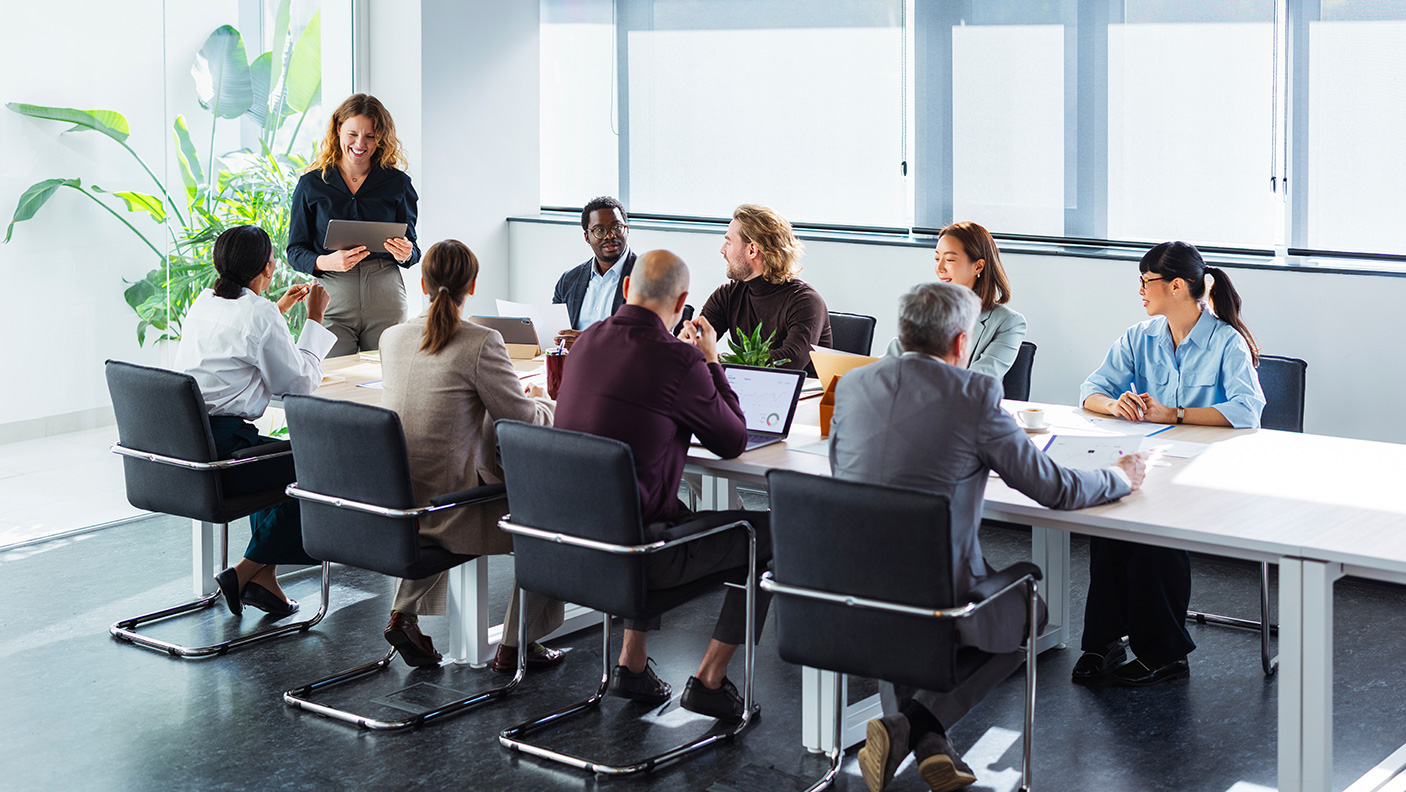 8 people sit around a board room table, with a female leader standing at the head of the table, presenting to the group