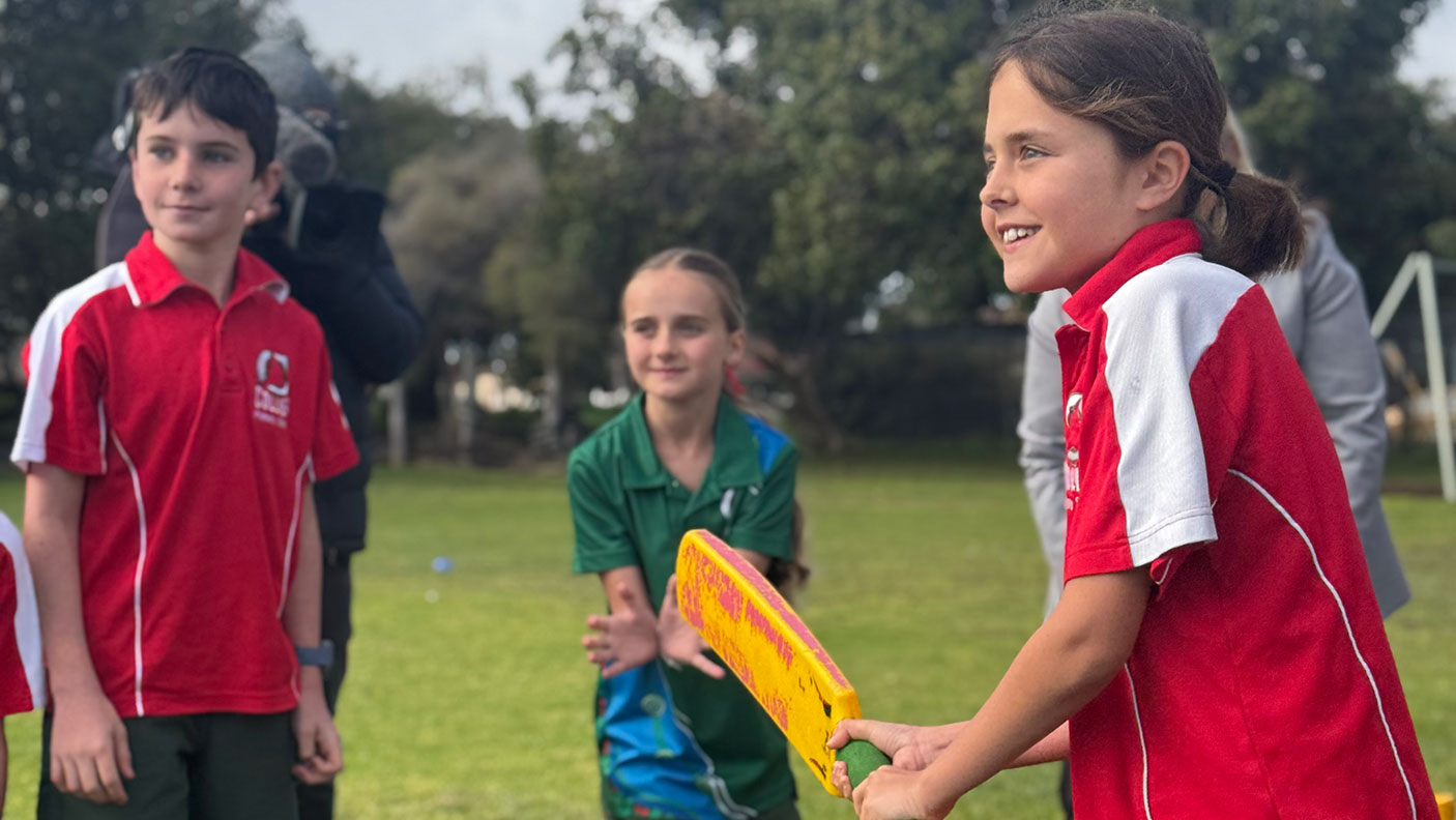 A primary school student holds a cricket bat, with two other students in the background