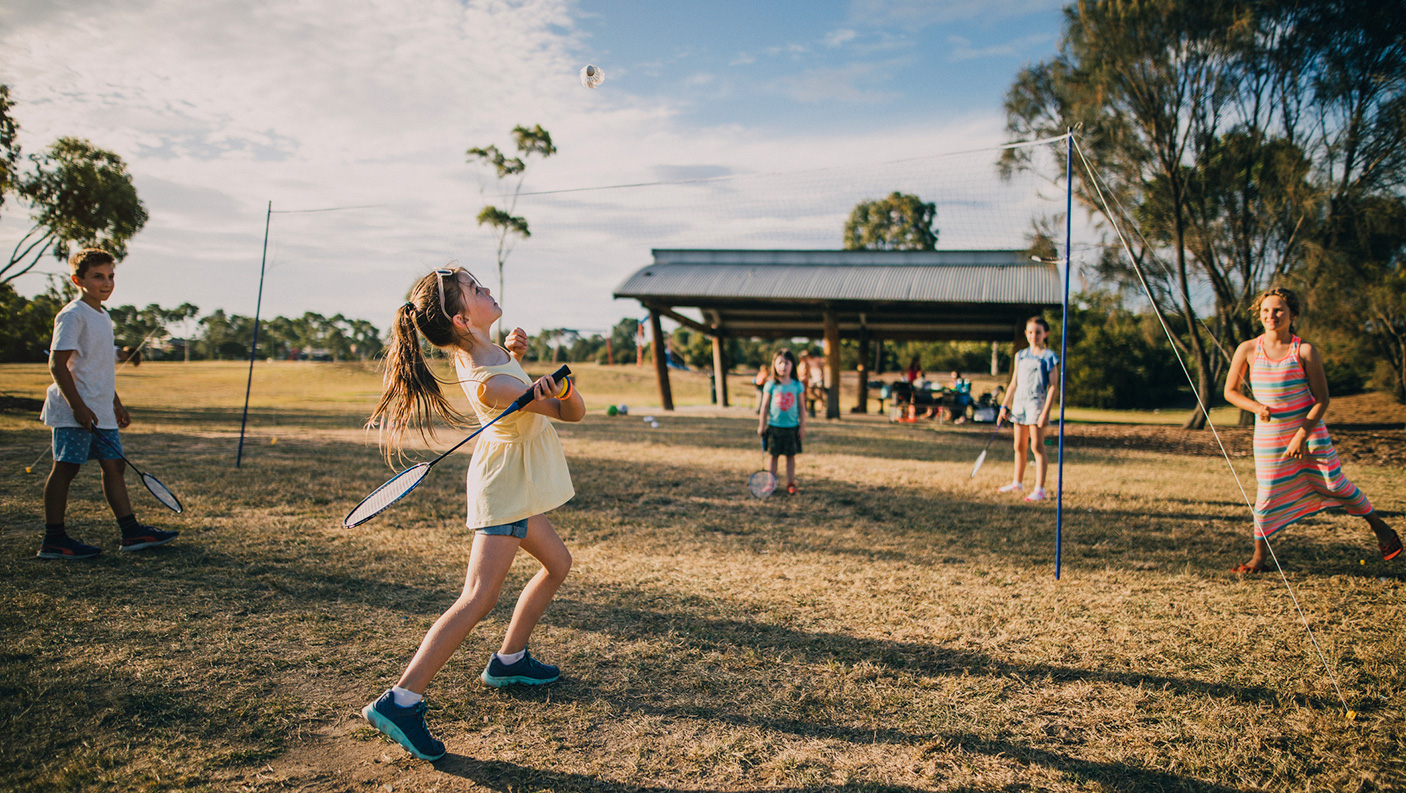 a small group of children playing badminton in a park, with a gazebo and gum trees in the background