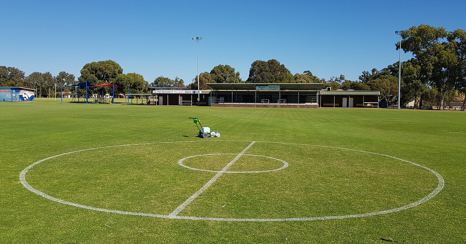 A grassed sports playing field with a clubroom and trees in the background