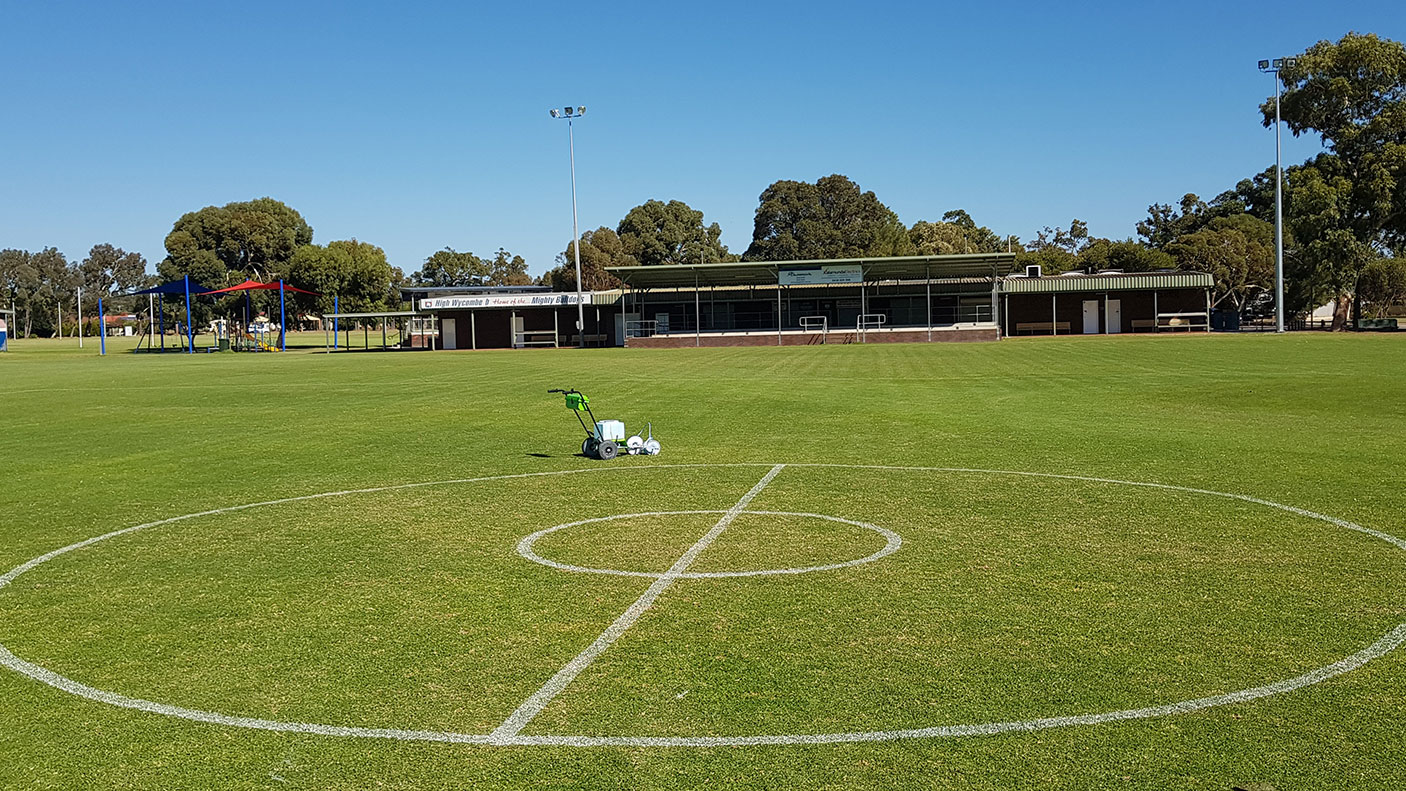 A grassyy sports playing field with a clubroom and trees in the background