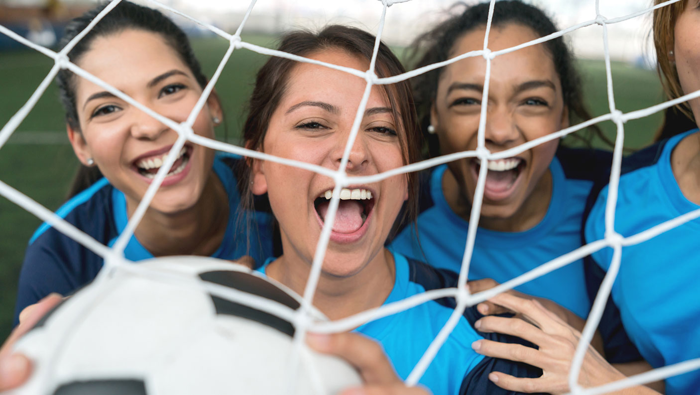 Women-football-web 4 women football players hold a football behind the goal nets