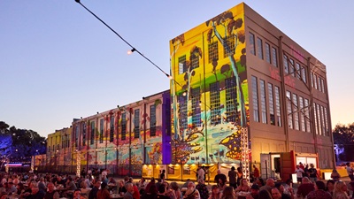 Crowd gathered outside a multi-storey building featuring a large colourful mural at dusk